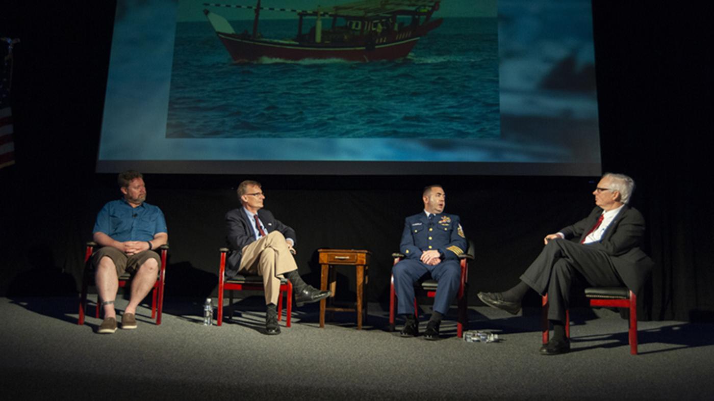 Four men seated speaking at a symposium