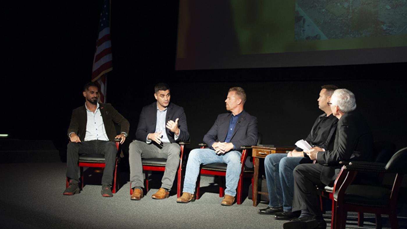 Five men seated speaking at a symposium