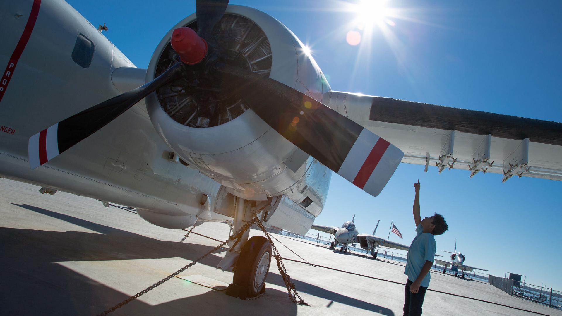 a small boy stands next to a large aircraft pointing to the wing above his head