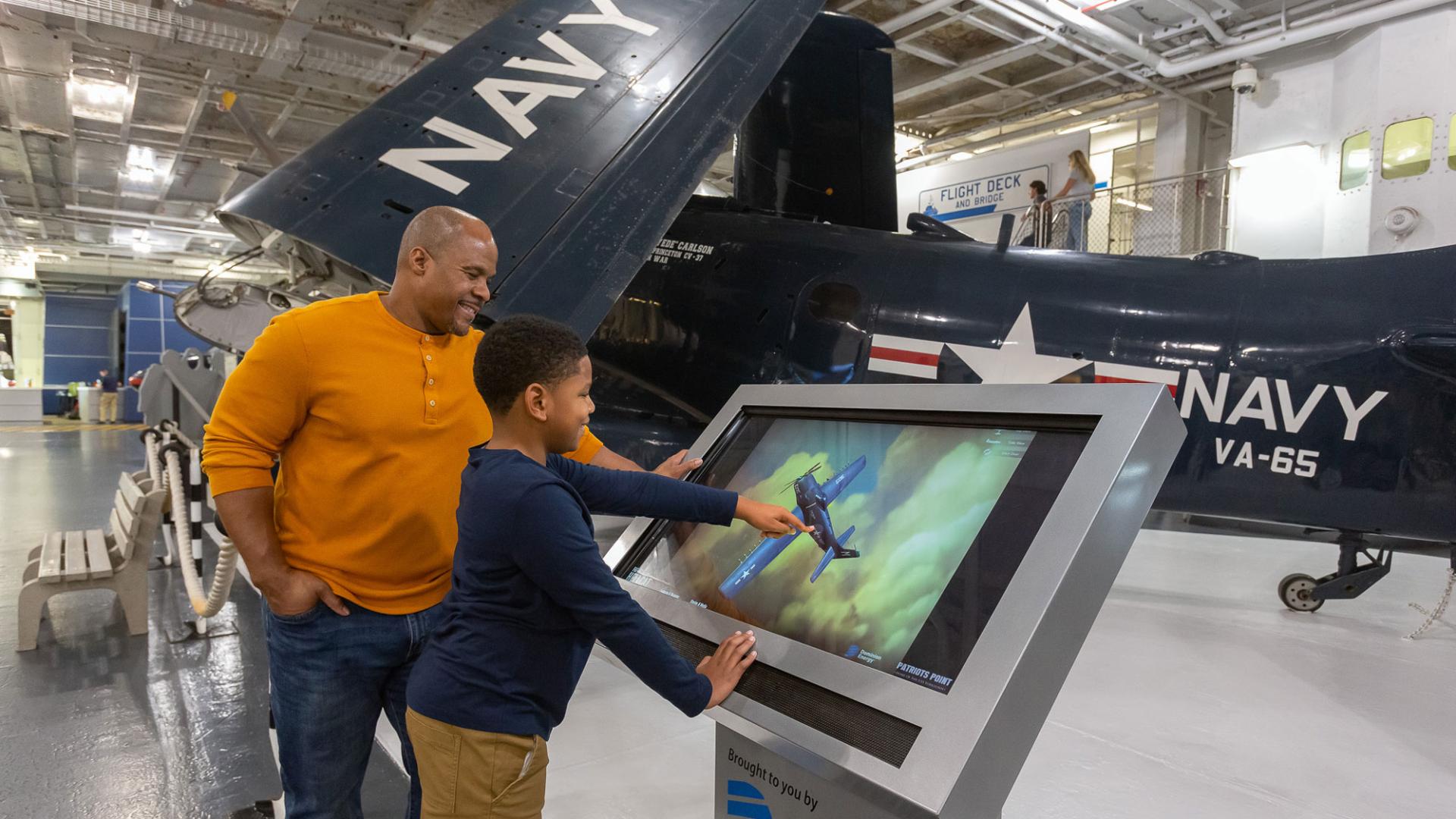 a father and son use a touch screen kiosk in front of a US Navy plane in a museum