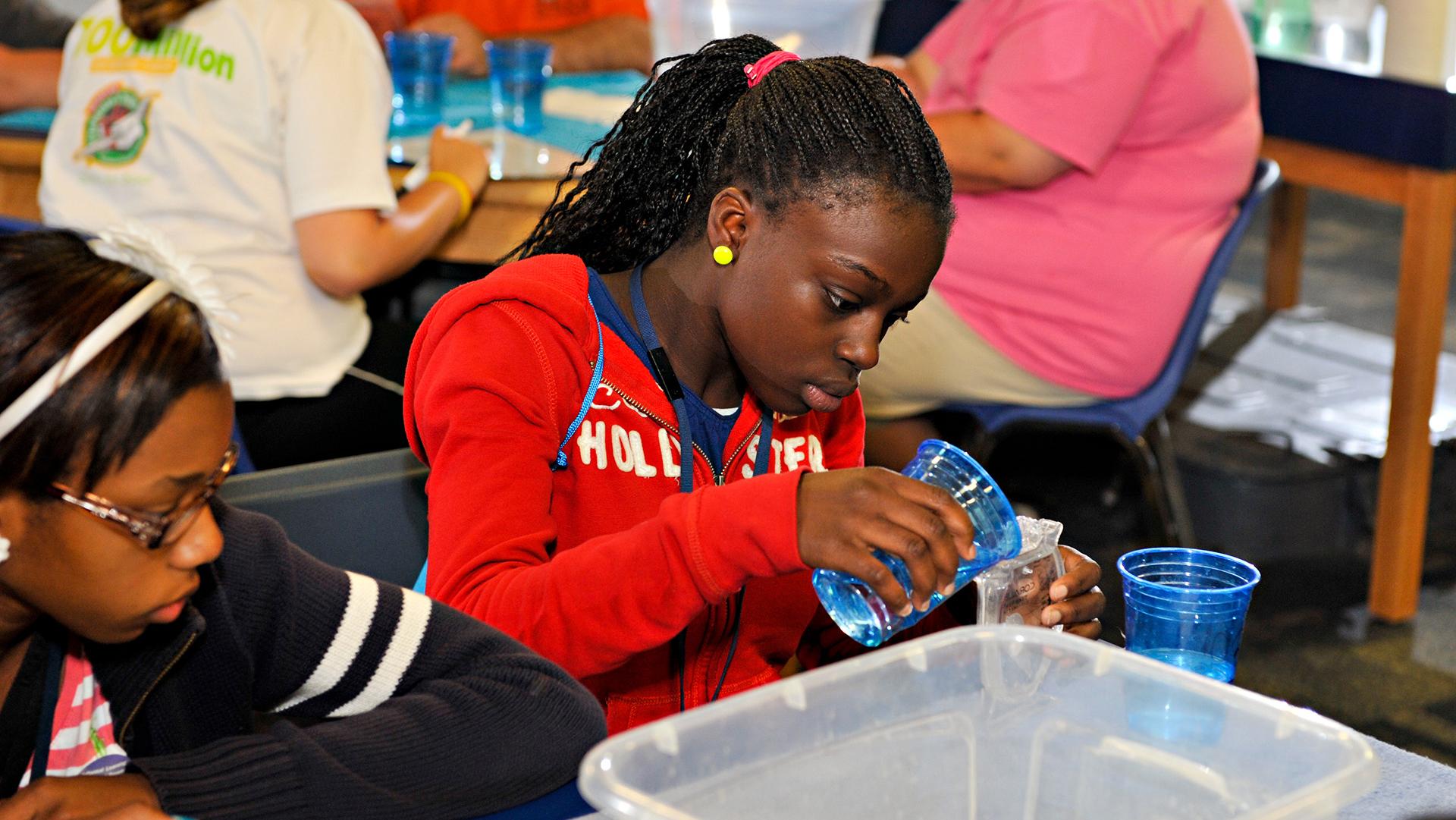 One girl watching another pour a liquid from one container to another