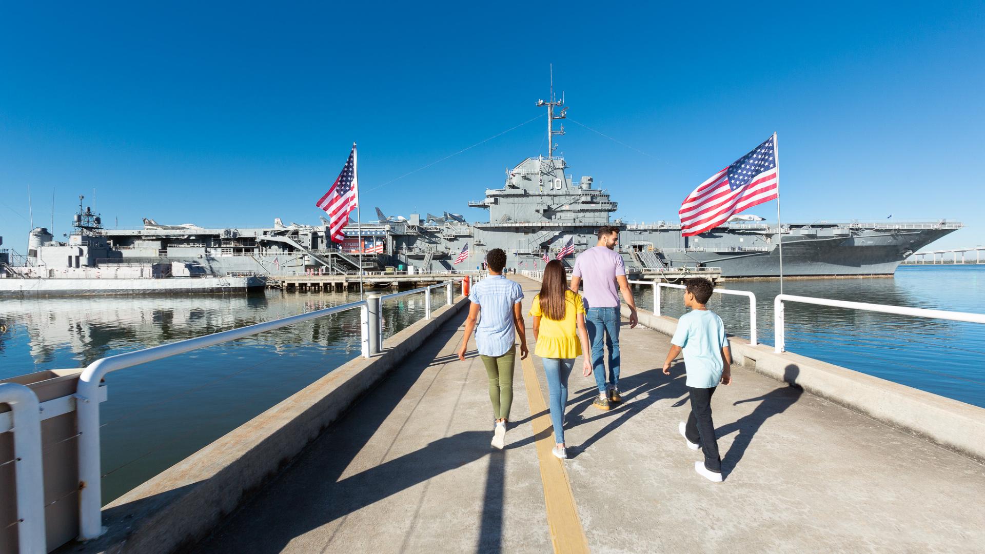 A family walks down the pathway toward the USS Yorktown aircraft carrier at Patriots Point, surrounded by American flags and calm waters. 