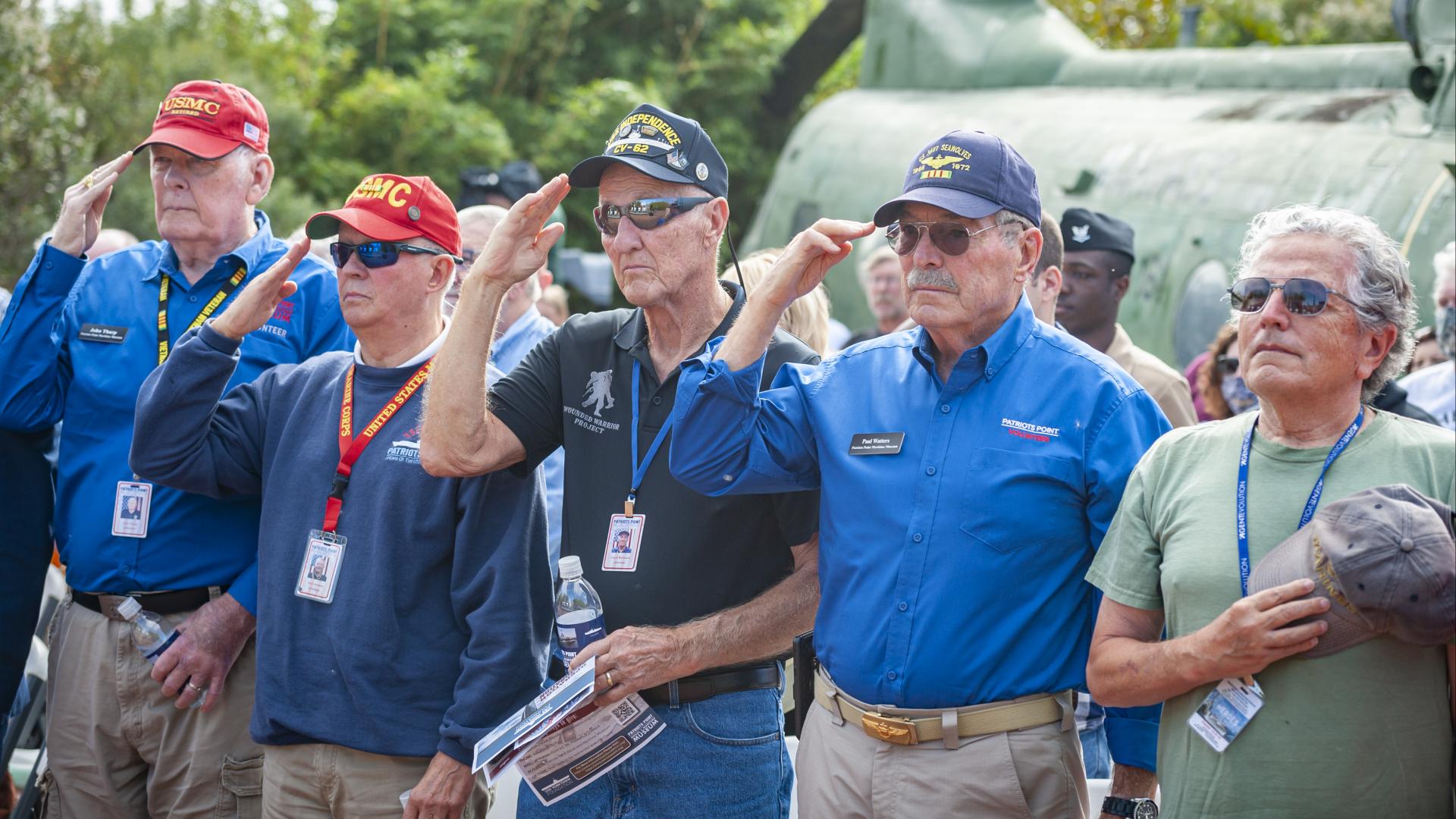 Group of veterans and volunteers at Patriots Point salute during a commemorative event in front of a military helicopter.