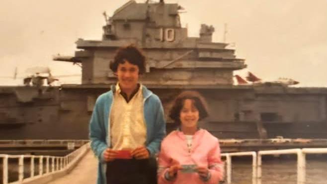 A boy in blue jacket and girl in pink shirt stand in front of the ship USS Yorktown with the number 10 on the side.