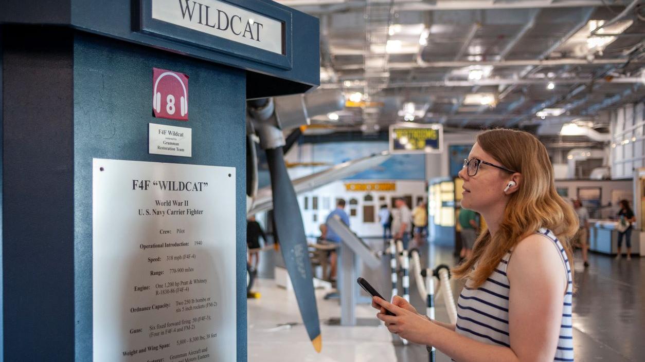 Woman standing in front of signage that explains the audio tour available for the F4F Wildcat exhibit