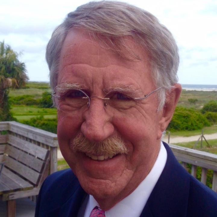 A man with salt and pepper hair, a mustache and glasses standing outside in a suit and tie
