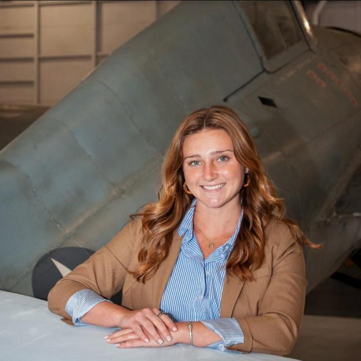 Headshot of Meaghan. Woman with long slightly curled brown hair wearing a brown suit jacket and a blue and white stripped shirt