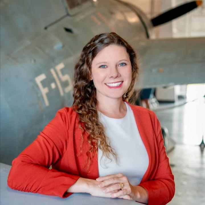 Headshot of Kayla Webber. Woman with long brown curly hair wearing an orange suit coat and a white shirt