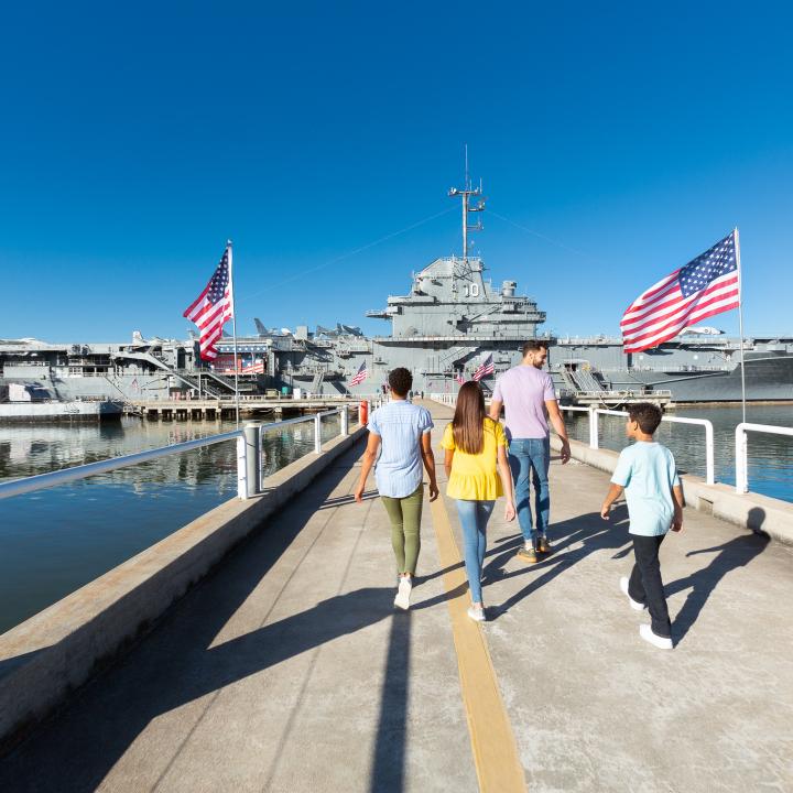 A family walks down the pathway toward the USS Yorktown aircraft carrier at Patriots Point, surrounded by American flags and calm waters. 