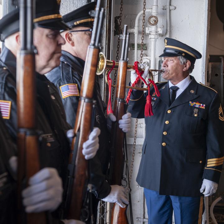 A solemn honor guard ceremony for National Pearl Harbor Remembrance Day aboard the USS Yorktown, with uniformed servicemen standing in formation holding rifles, while a bugler in military dress plays a ceremonial tune.