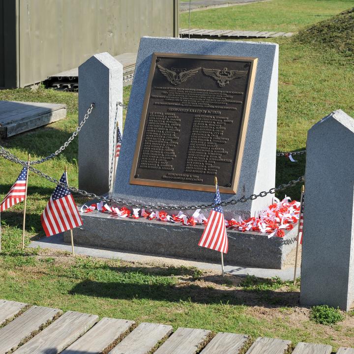 Memorial monument featuring a plaque engraved with the names of servicemembers killed in action, surrounded by small American flags and red, white, and blue decorations. 