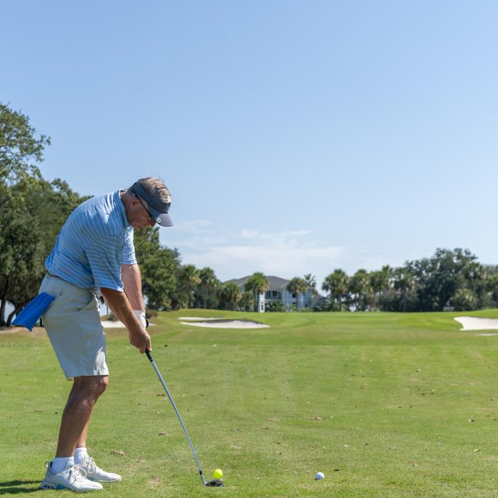 A golfer prepares to take a swing on a sunny day, standing on a well-maintained golf course surrounded by trees and distant homes.