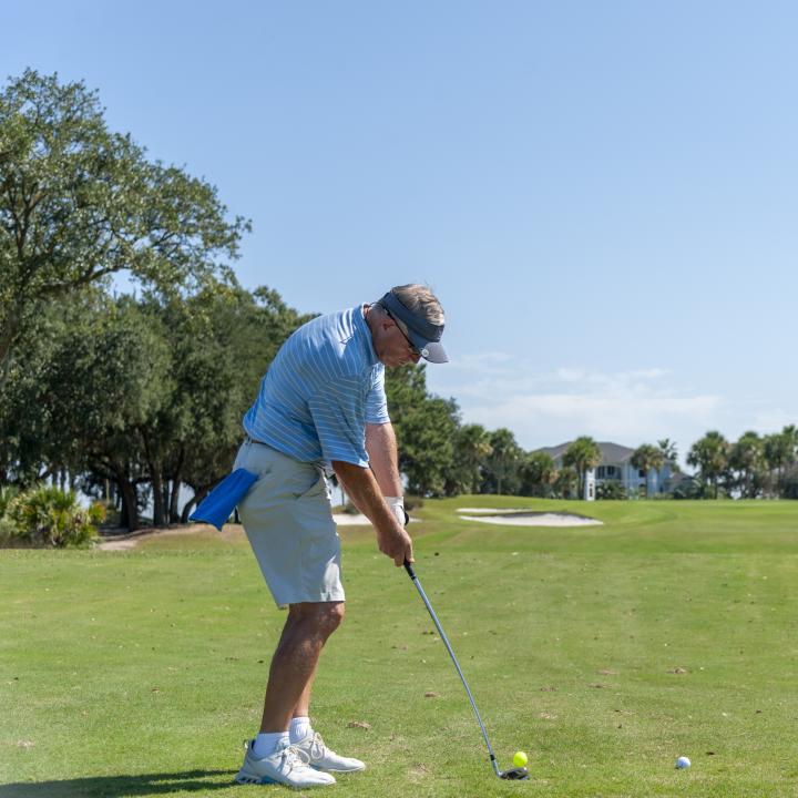 A golfer prepares to take a swing on a sunny day, standing on a well-maintained golf course surrounded by trees and distant homes.