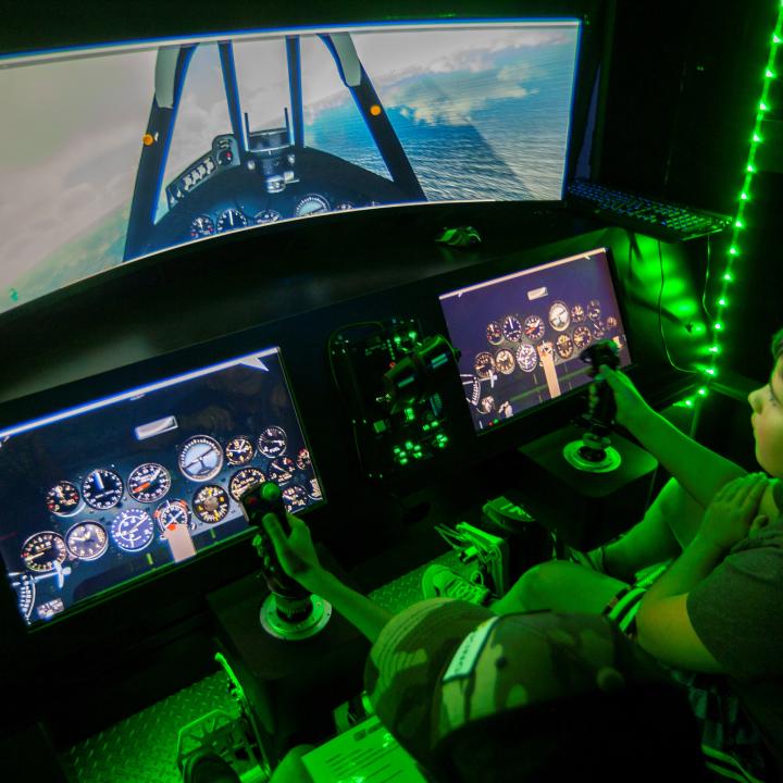 Child uses handheld joystick to virtually fly military aircraft illuminated by green lights.