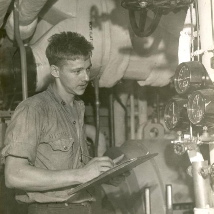 A young man downstairs in the engine room taking notes on the equipment.
