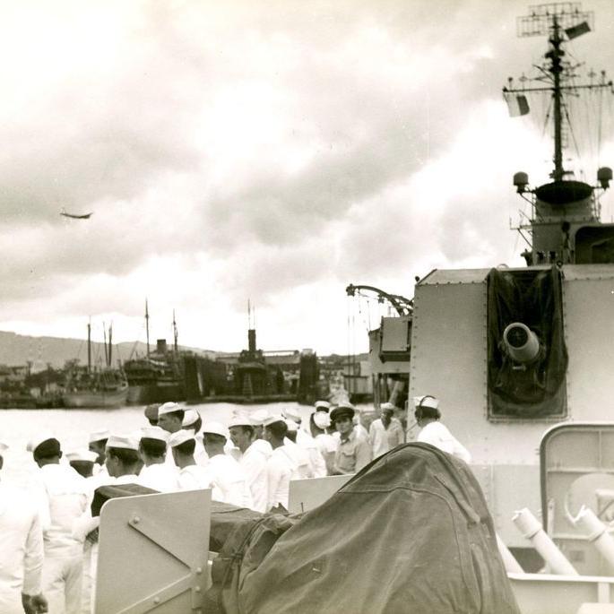 Men in white sailors' uniforms getting ready to disembark at Pearl Harbor. 5'-gun turret is facing the camera and aircraft is flying in background.