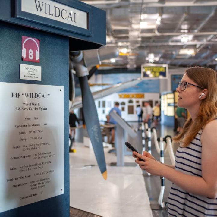 Woman standing in front of signage that explains the audio tour available for the F4F Wildcat exhibit