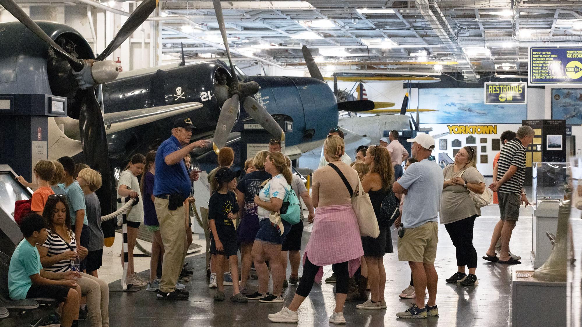 a museum volunteer speaks to a crowd of museum visitors in front of several vintage aircraft