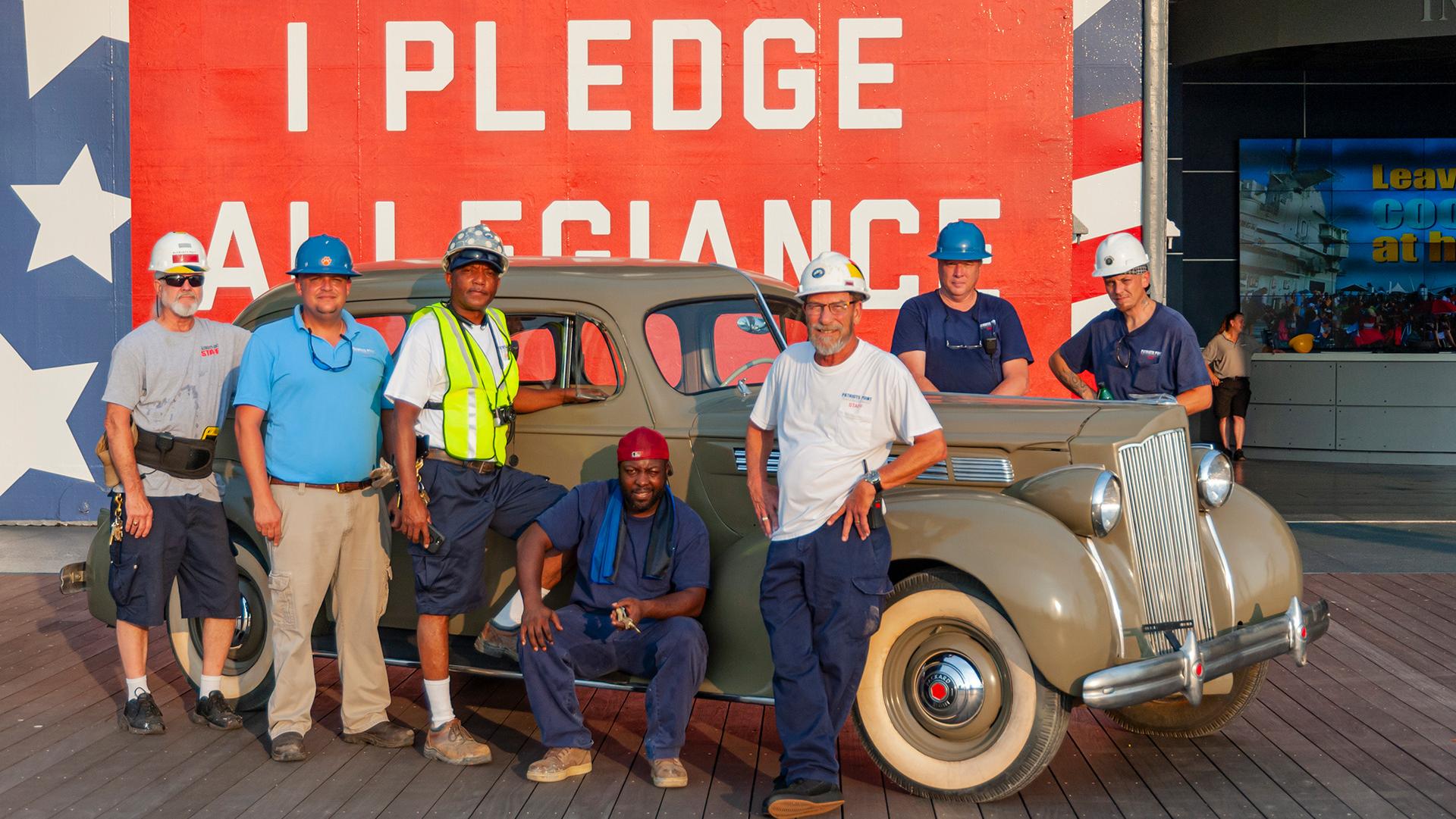 Seven individual in hard hats stand around an older car