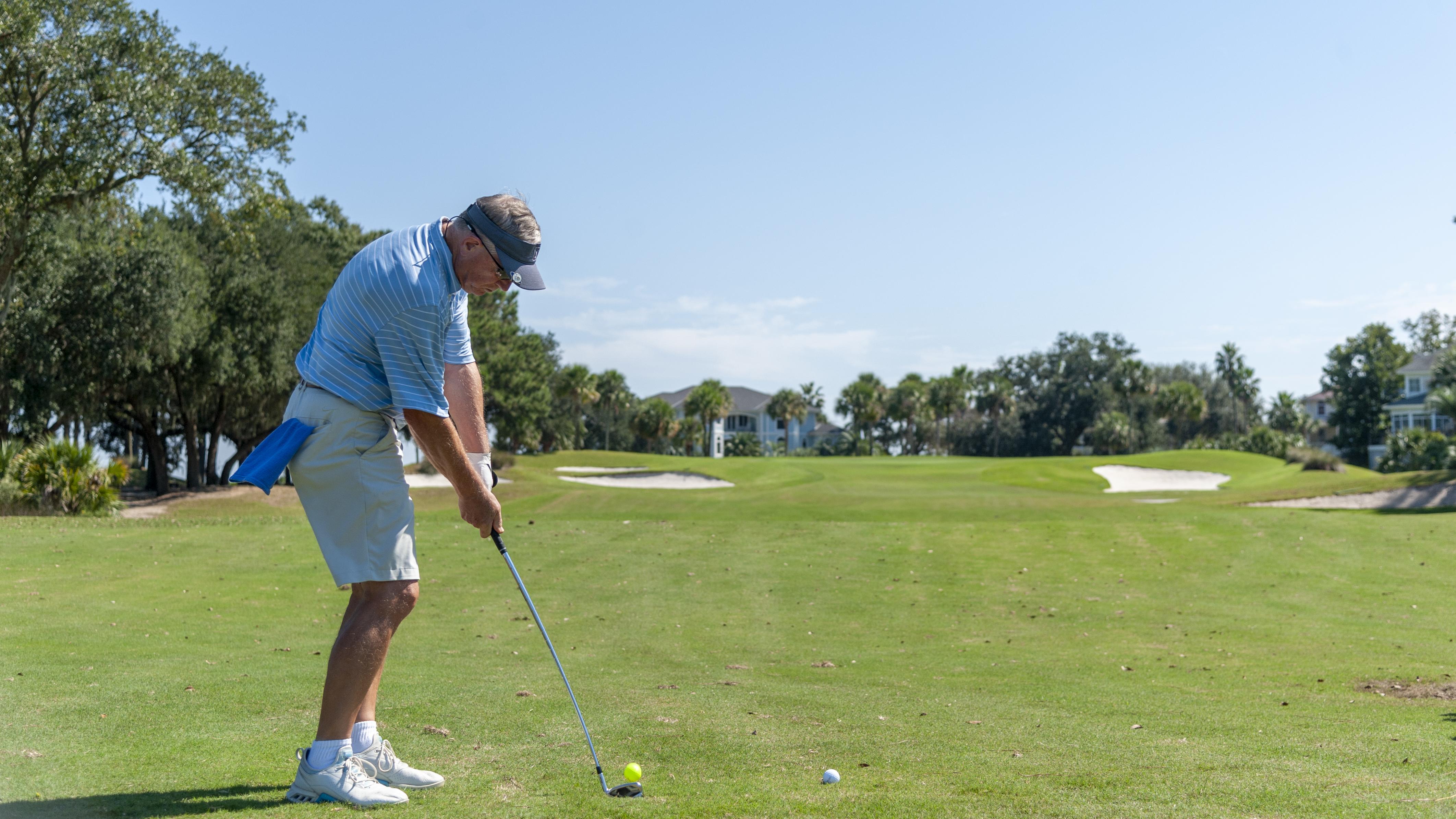 A golfer prepares to take a swing on a sunny day, standing on a well-maintained golf course surrounded by trees and distant homes.
