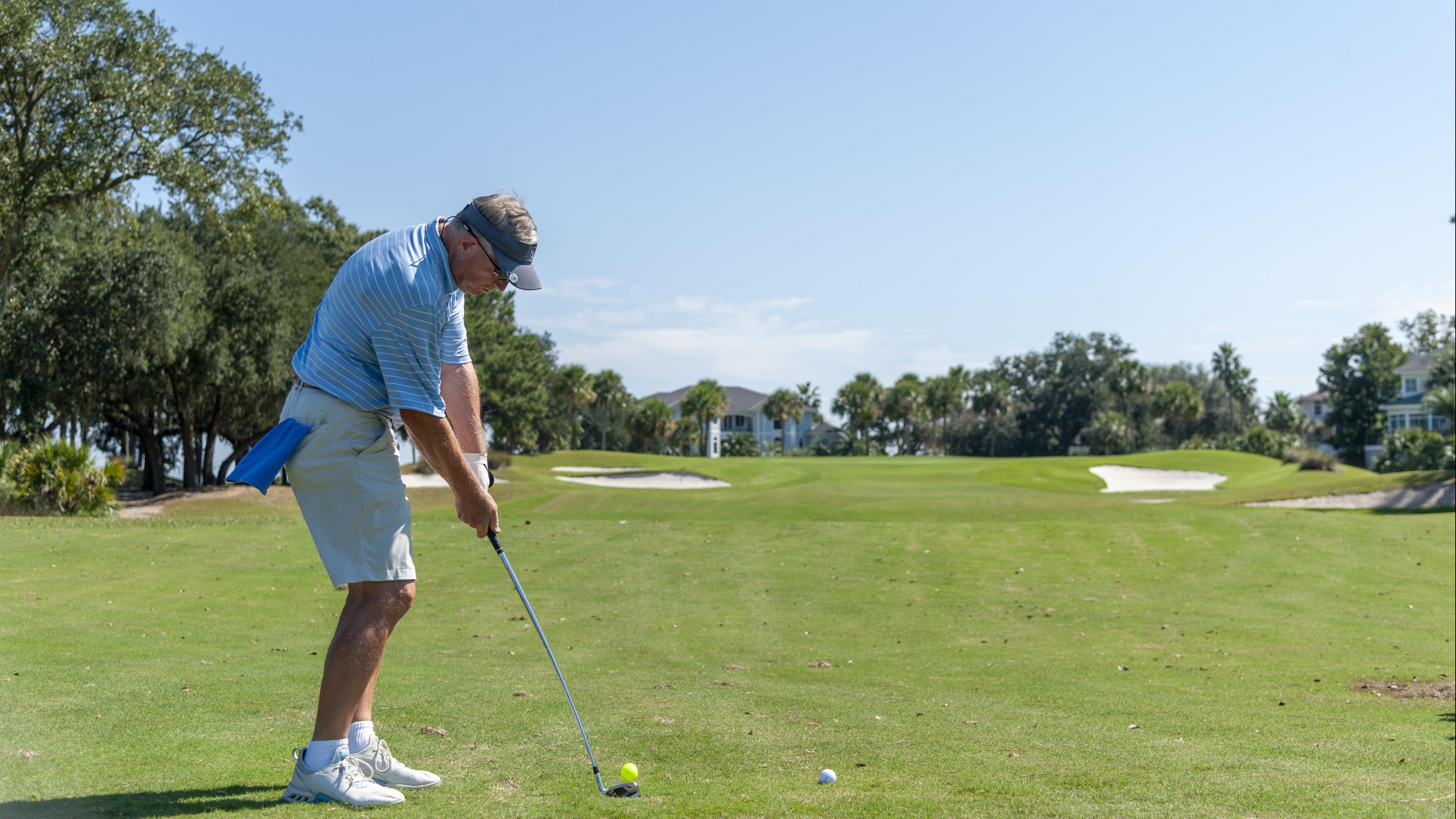 A golfer prepares to take a swing on a sunny day, standing on a well-maintained golf course surrounded by trees and distant homes.