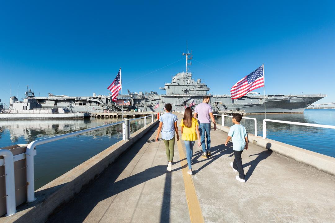 A family walks down the pathway toward the USS Yorktown aircraft carrier at Patriots Point, surrounded by American flags and calm waters. 