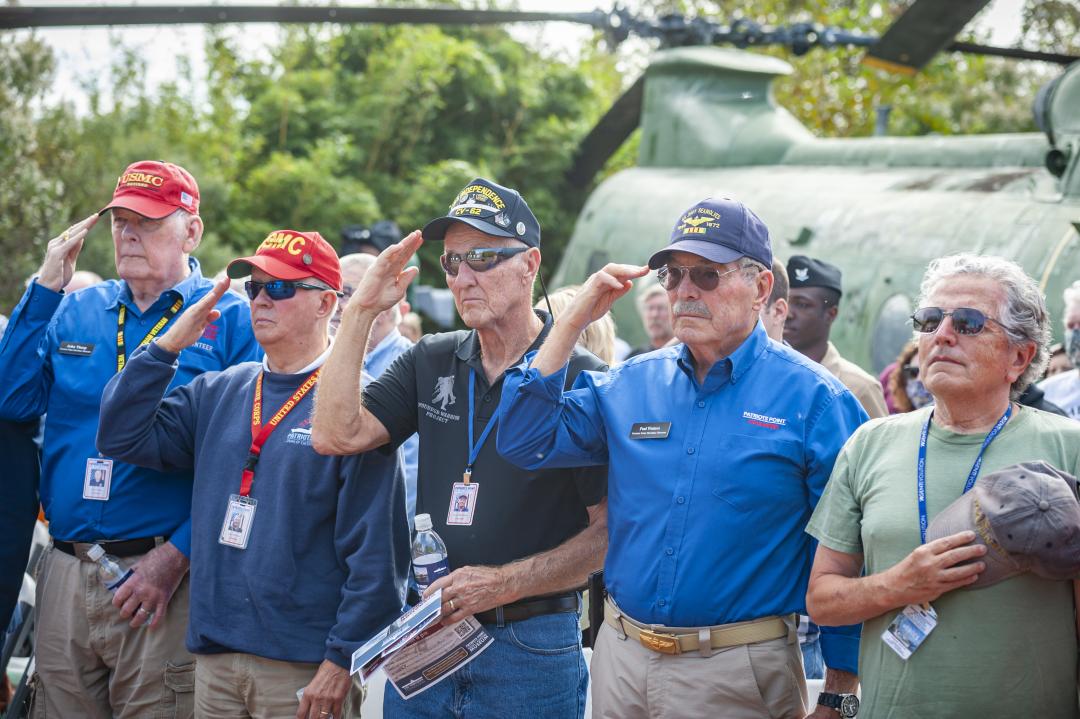 Group of veterans and volunteers at Patriots Point salute during a commemorative event in front of a military helicopter.