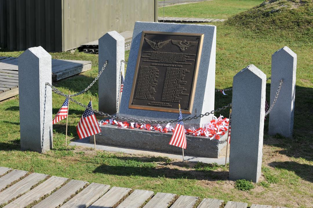 Memorial monument featuring a plaque engraved with the names of servicemembers killed in action, surrounded by small American flags and red, white, and blue decorations. 