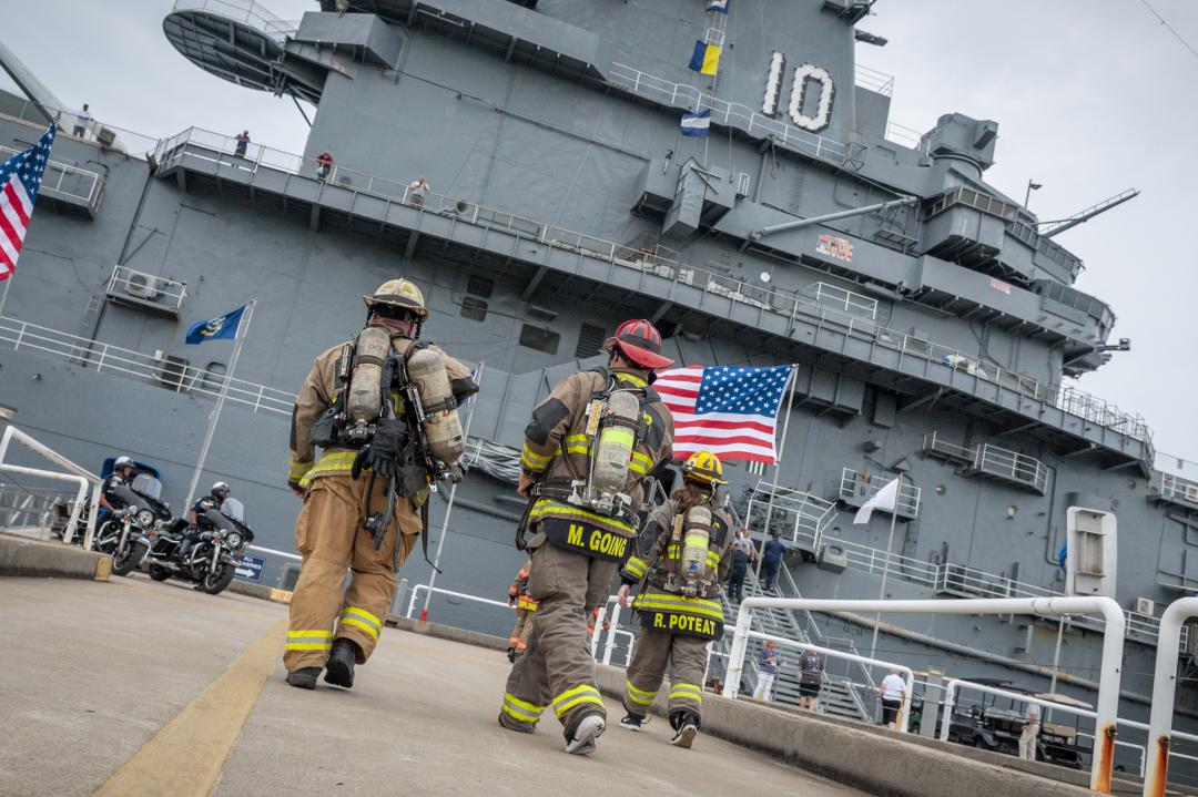 Firefighters in full gear walk toward the USS Yorktown, with an American flag prominently displayed.