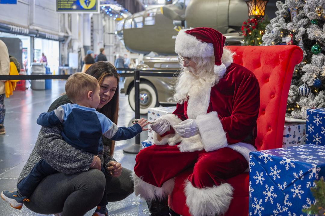 A young child and their parent interact with Santa Claus, who is seated in a red chair and handing the child a candy cane, surrounded by festive holiday decorations and gifts.