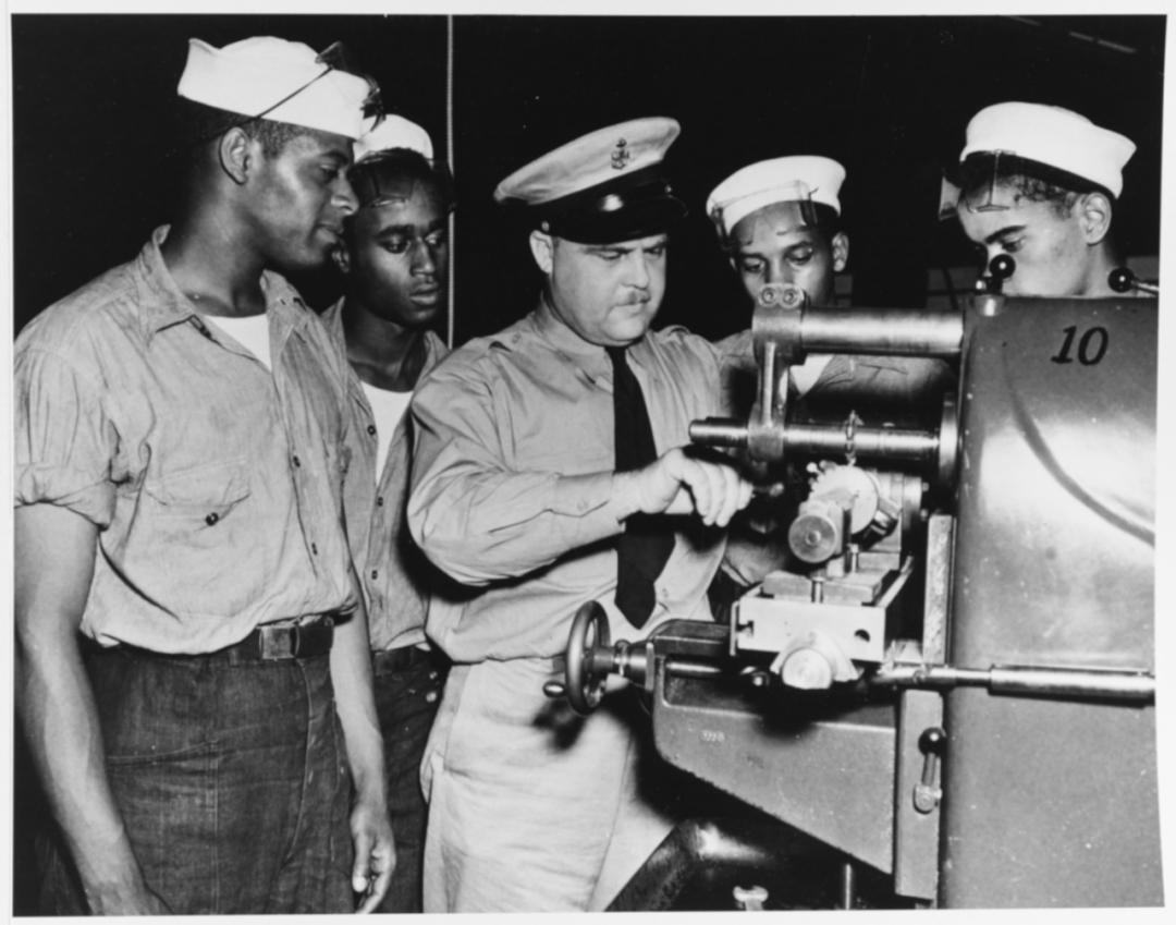 Four black sailors standing over a piece of ship machinery, being taught by a white Chief Petty Officer. 