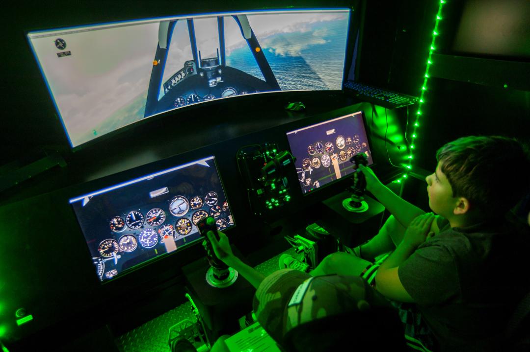 Child uses handheld joystick to virtually fly military aircraft illuminated by green lights.