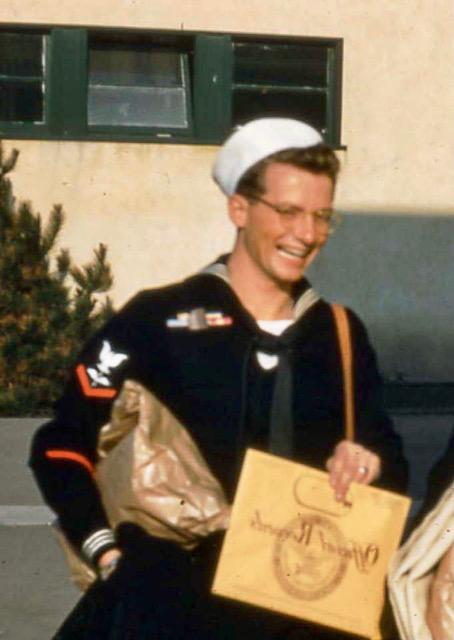 Young sailor dressed in blue uniform stands with yellow colored paperwork and a plastic bag. He has glasses. 