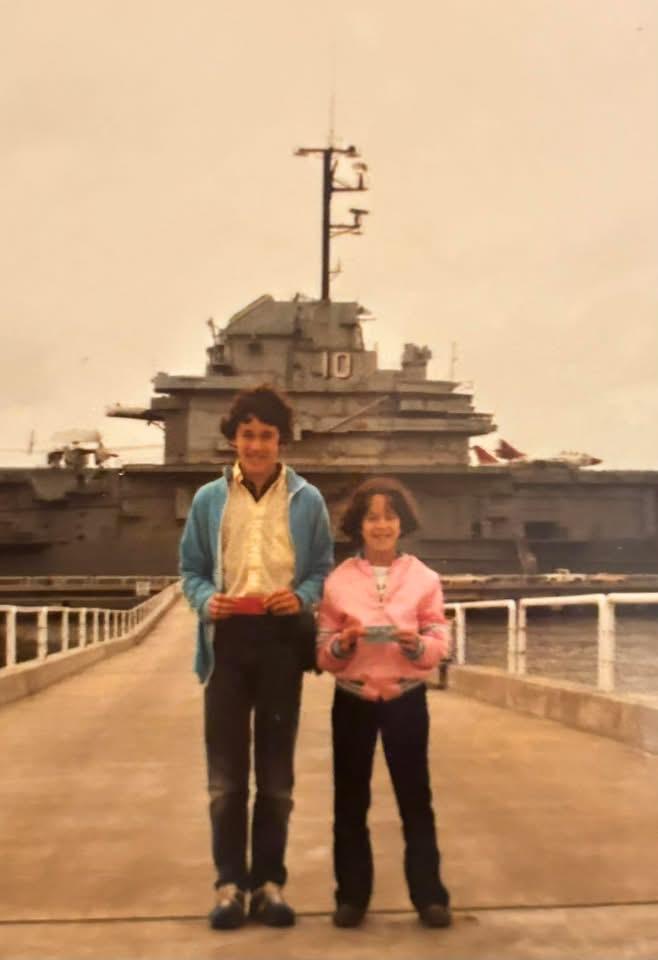 A boy in blue jacket and girl in pink shirt stand in front of the ship USS Yorktown with the number 10 on the side.