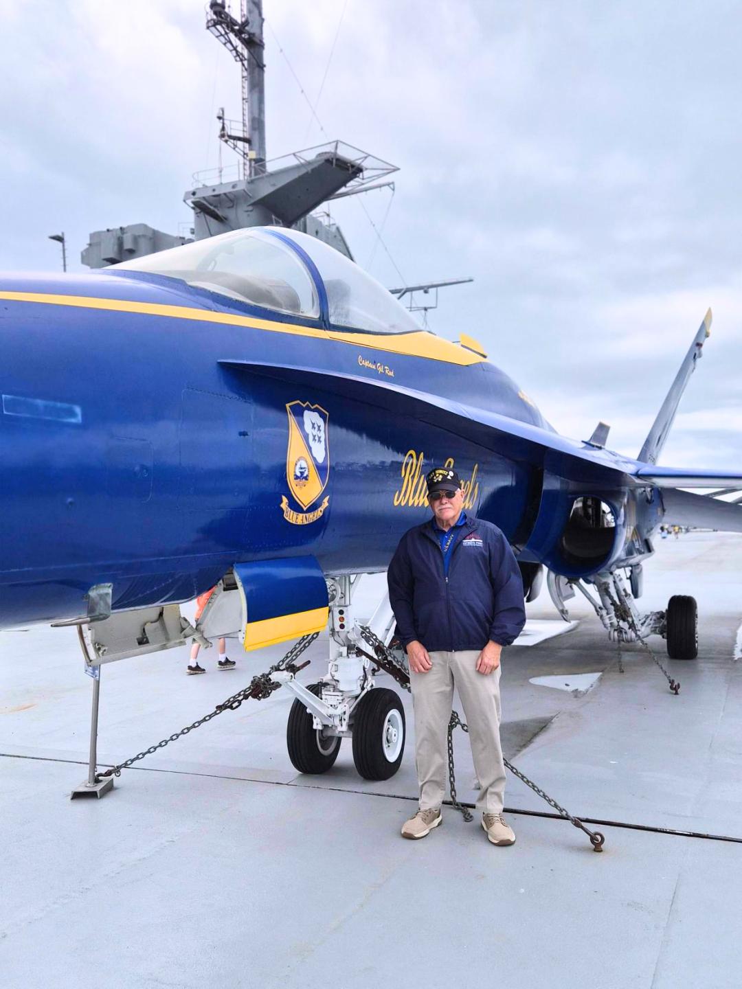 Painted aircraft with 'Blue Angel' on aircraft carrier flight deck and veteran Tom Simons in front of it.