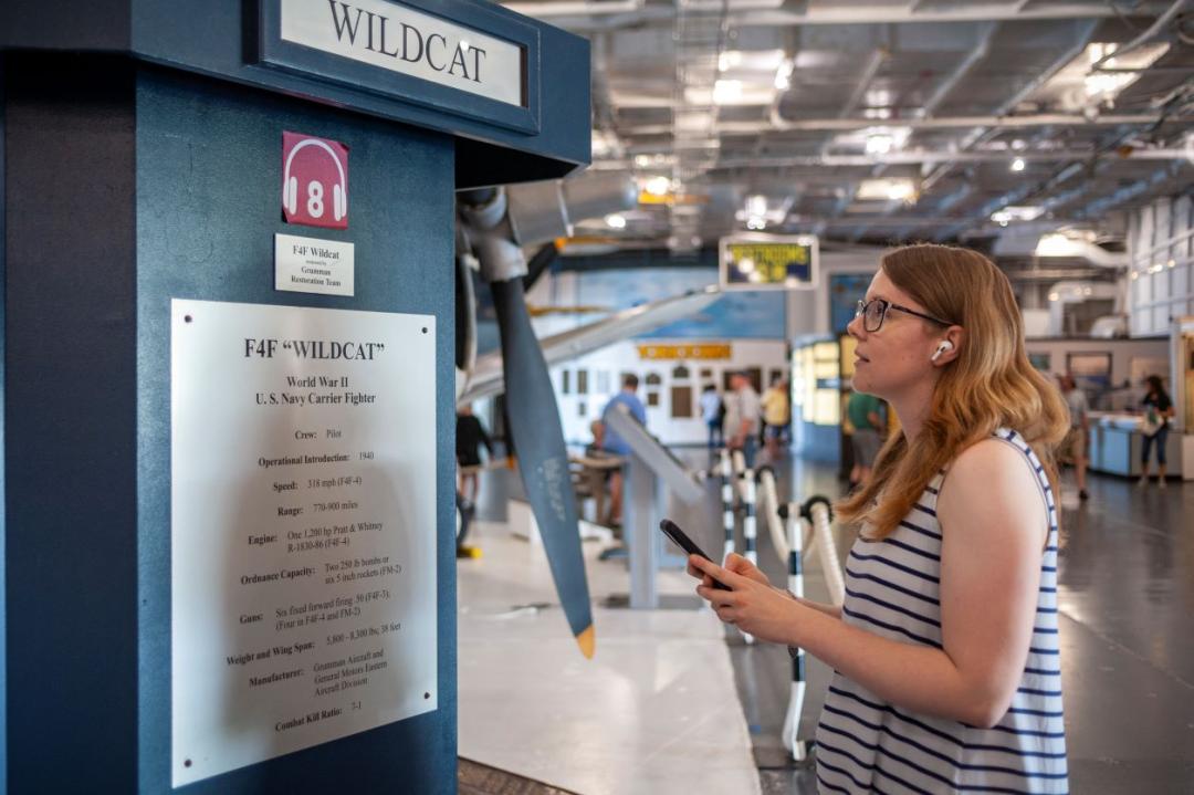 Woman standing in front of signage that explains the audio tour available for the F4F Wildcat exhibit